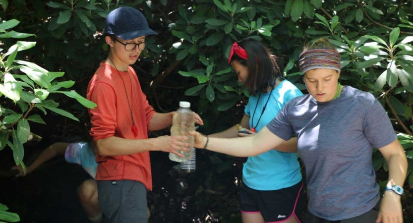 Two students pass a bottle of water between them. There are other students and green leaves in the background. 